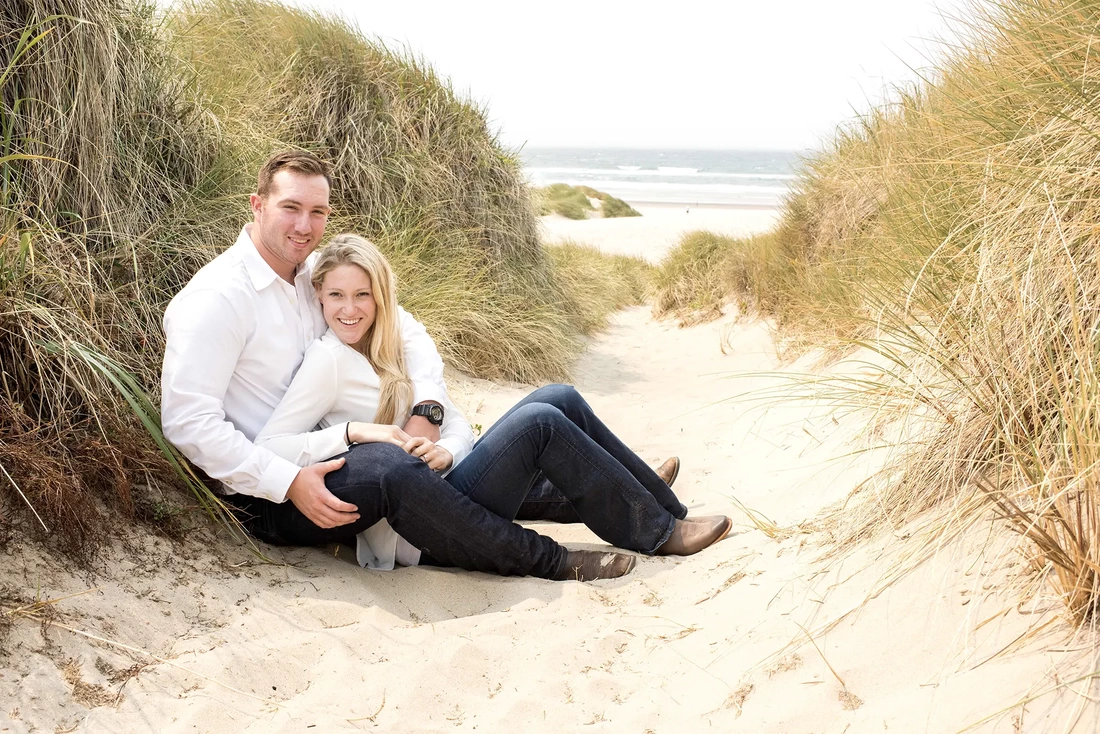 Oregon Coast Engagement Photos With Modern Art Photograph, a deep path is carved between the sand dunes a couple rests against the wall of the dune, they look to the camera, the ocean is in the distance