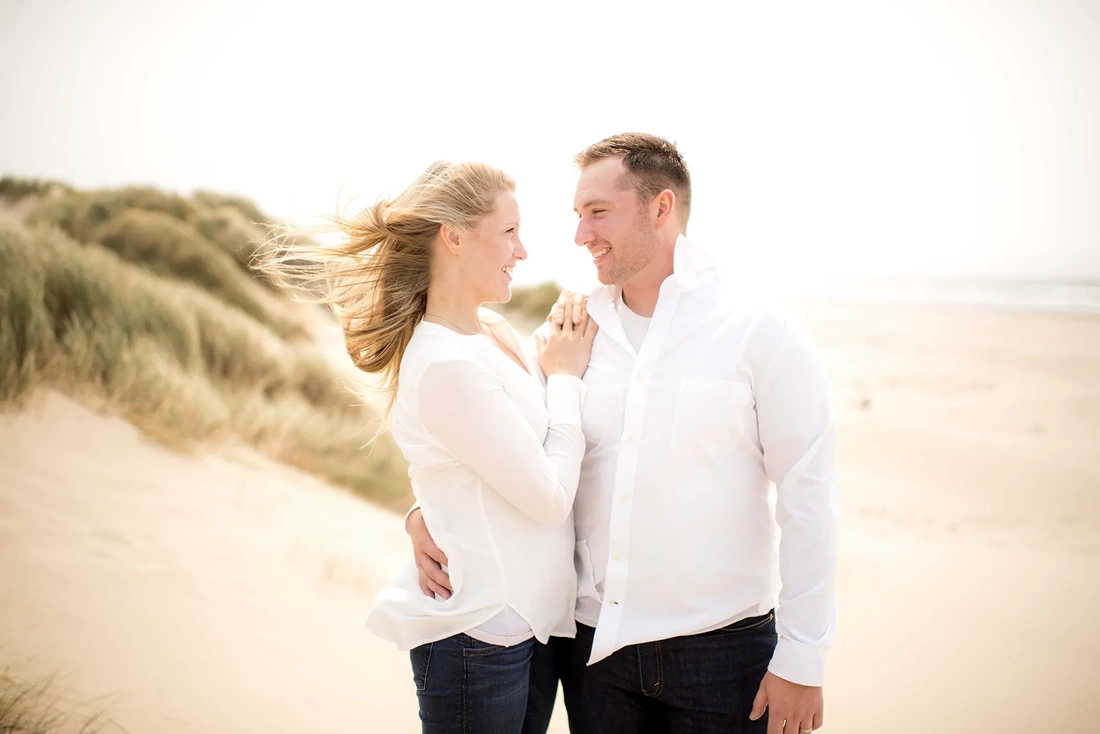 Oregon Coast Engagement Photos With Modern Art Photograph a man and woman stand in amber light on the beach and look into each others eyes, the wind blows the woman's hair 