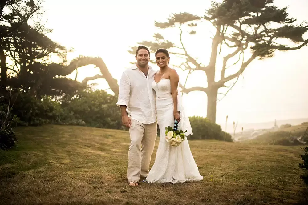 Photographers Portland at Wedding On The Beach, this bride and groom stand together on the grasses near their wedding venue, twisted trees and sunset light through the fog over the ocean light the scene 
