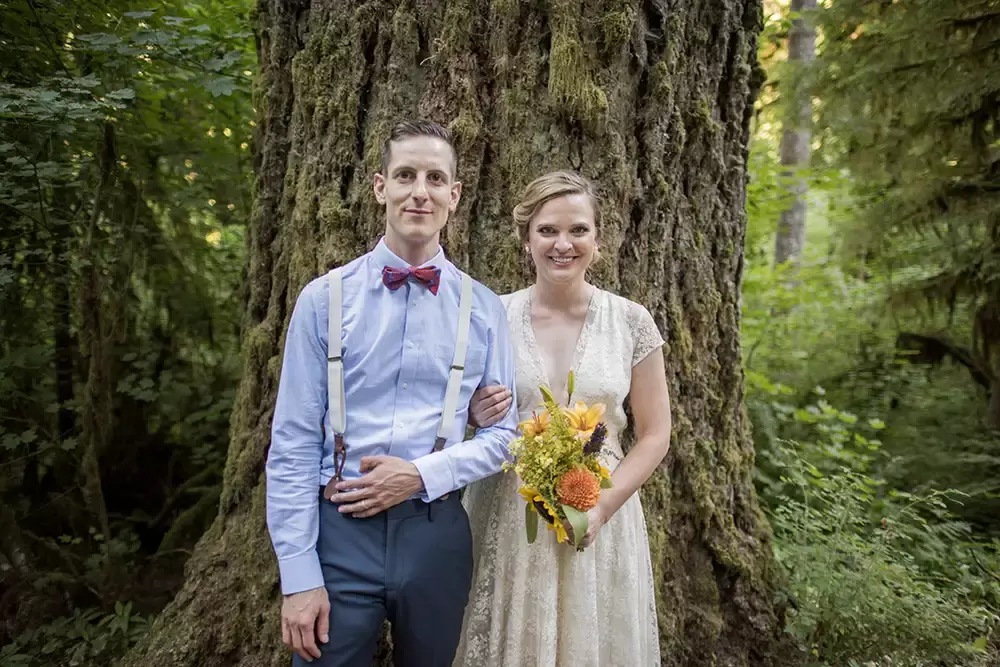 Photographers Portland at Silver Falls Weddings, A bride and groom in semi formal attire stand arm in arm agains an old growth fir tree. The forest falls out of focus behind them. The texture of the forest is thick and heavy. The couple is sharply dressed and beaming with excitement for the wedding ahead. 