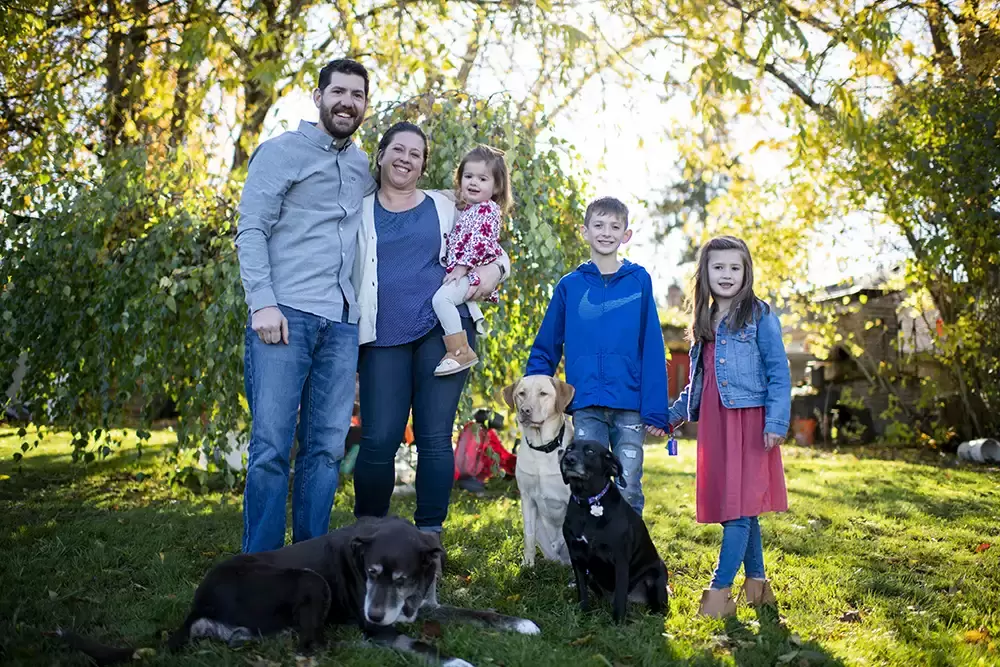 Portland Family Photographer Robert Knapp takes a photo of a family together in their back yard with three dogs. The autumn light shines behind them toward the camera 