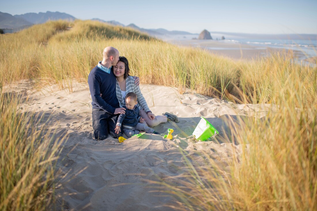 A husband kisses his wife on the top of the head while she looks at the camera. Their child plays in the sand with toys. They are sitting in the sand surrounded by tall grasses with a view of the oregon coast behind them. The light is early morning, the shadows are long .