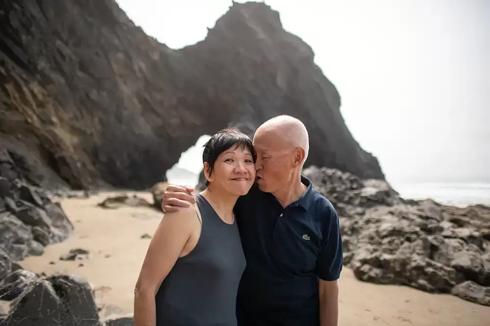 ​grandpa kisses grandma on the cheek. They both smile. in the background a great arch grows from the cliff to the ocean Portland ​Family Photographer Robert Knapp - Book Today!