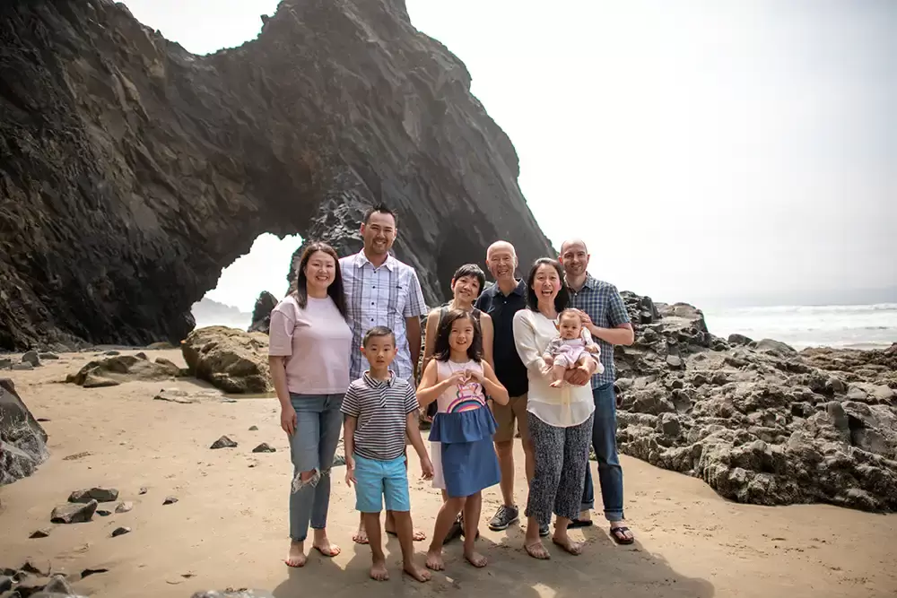 a close up of a family smiling at the camera with the ocean and a stone arch in the background. Portland ​Family Photographer Robert Knapp - Book Today!