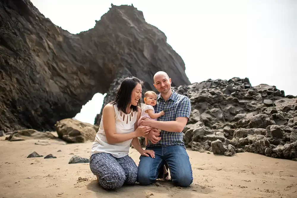 mom smiles at baby and daddy on the beach for a family photo near some interesting rock formation Portland ​Family Photographer Robert Knapp - Book Today!