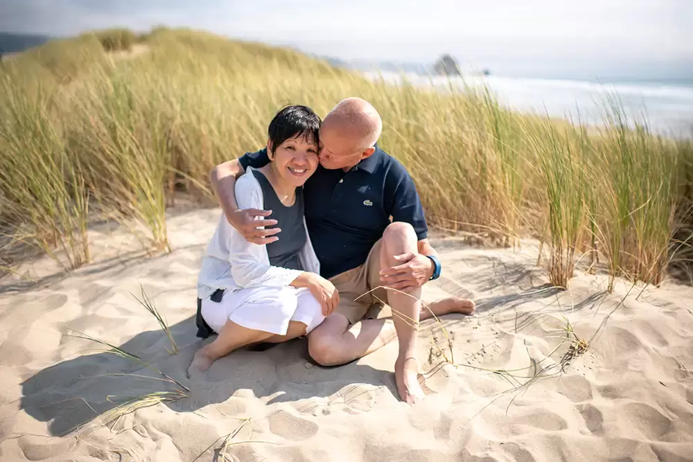 Grandpa kisses grandma on the cheek. They're both sitting in the sand. The ocean is in the distance. Portland ​Family Photographer Robert Knapp - Book Today!b​Family Photographer Robert Knapp in Portland - Book Today!