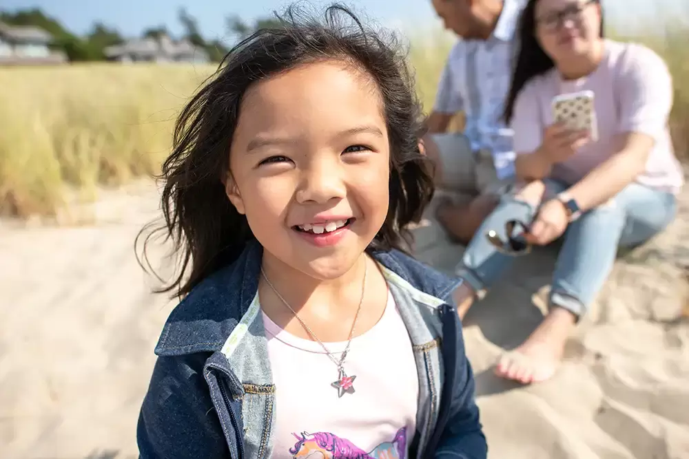 A little girl smiles to the camera in the background on the beach​Family Photographer Robert Knapp in Portland - Book Today!