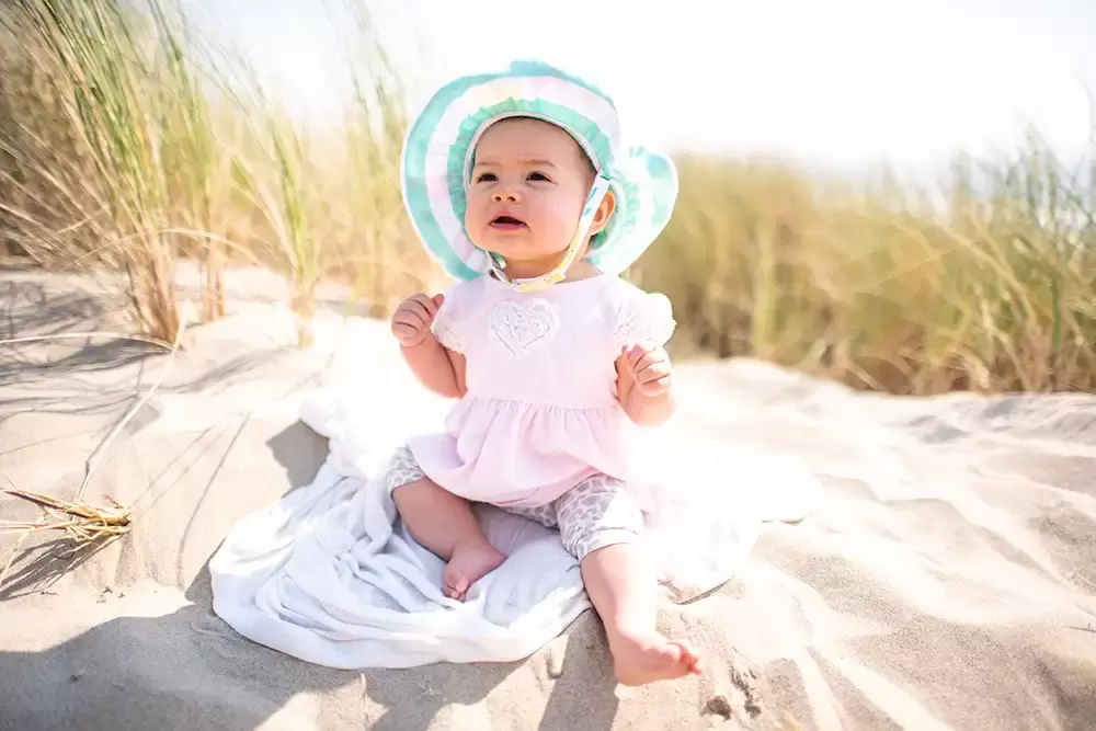 baby looks up to mama. baby sits in the sand on a towel. Sea grasses are around. ​Family Photographer Robert Knapp in Portland - Book Today!
