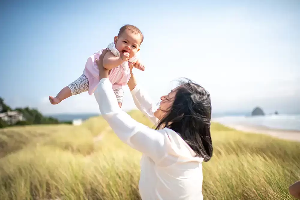 mother holds baby up to the air. grassy dunes roll in the distance Portland ​Family Photographer Robert Knapp - Book Today! ​Family Photographer Robert Knapp in Portland - Book Today!