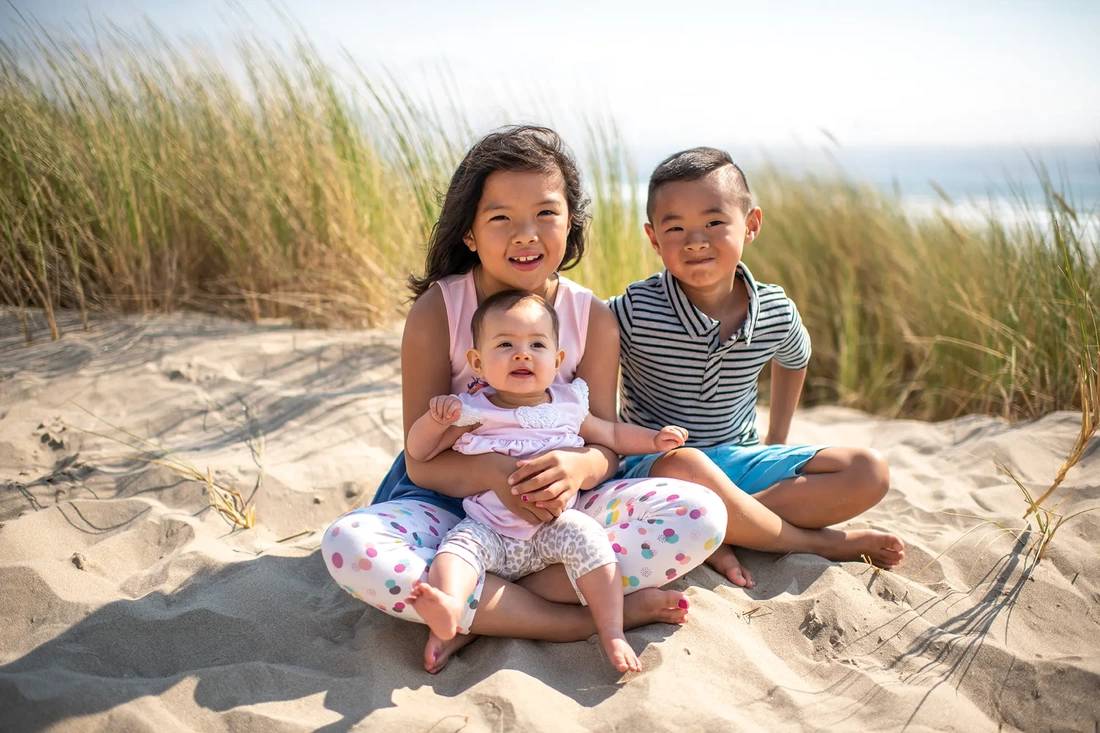 small children sit together and smile in the sand. Sea grasses and ocean in the background Portland ​Family Photographer Robert Knapp - Book Today!