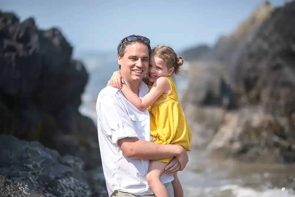 father and daughter snuggle with the ocean and the waves behind them   Family Pictures Beach Theme with Portland Family Photographer Robert Knapp