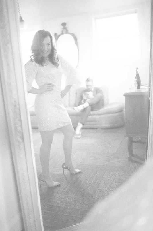 Play photograph through a mirror, the bride poses and smiles, and is kind of pushing her hips out in the background out of focus. The groom looks to his phone.