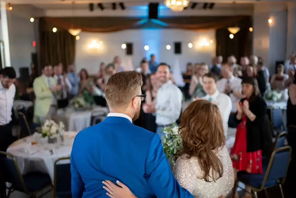 The bride and groom address their guests at the reception. The photograph is from the perspective behind an above the bride and groom. The back of their heads are in focus. All of the faces of the guests are looking through the camera, and the bride and groom they are on the f.