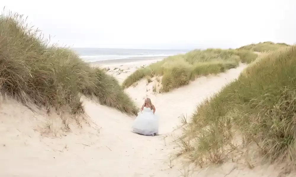 Photographers Portland at Bridal Photo shoot in the dunes, A woman in a white wedding dress climbs a large sand dune covered with sea grasses. the sand and grass split into paths around her. she looks to the interesting view. the ocean is in the distance
