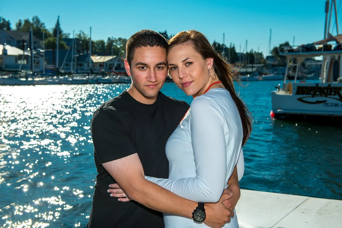 Engagement photo, a couple stands on the dock cheek to cheek looking at the camera. Sailboats are moored in the background