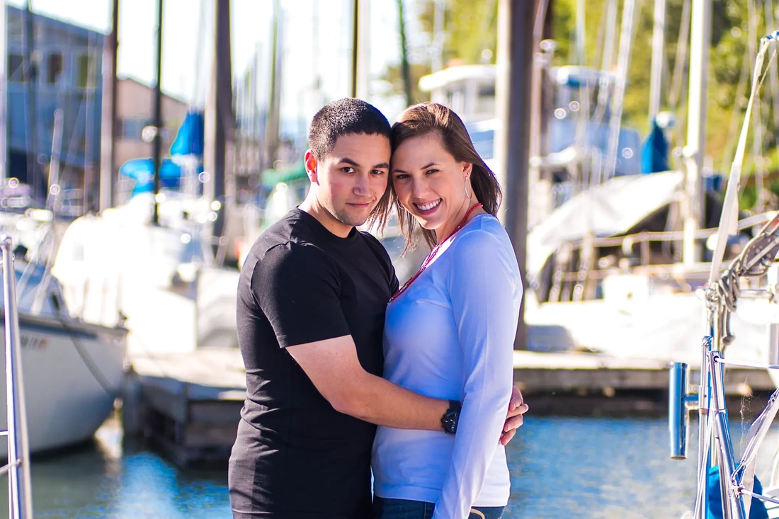A couple stands on the dock, holding each other they stand cheek to cheek with sailboats moored in the background
