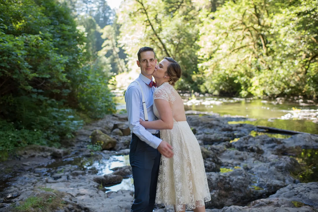 Silver Falls Weddings with Modern Art Photograph Standing along a riverbank in the shade, a bride kisses a groom on the cheek. The bride has her eyes closed, the groom looks to the camera with a smile at Silver Falls Weddings with Modern Art Photograph