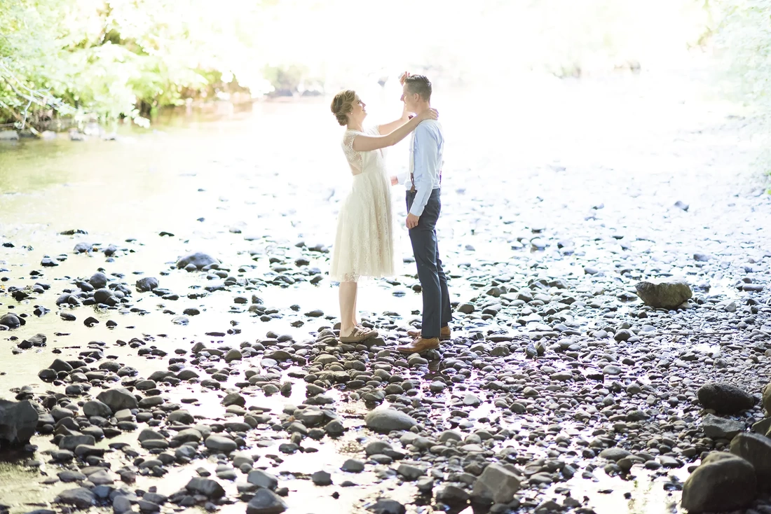 Silver Falls Weddings with Modern Art Photograph, a bride helps the groom with his collar standing on the stones of a river bank. 