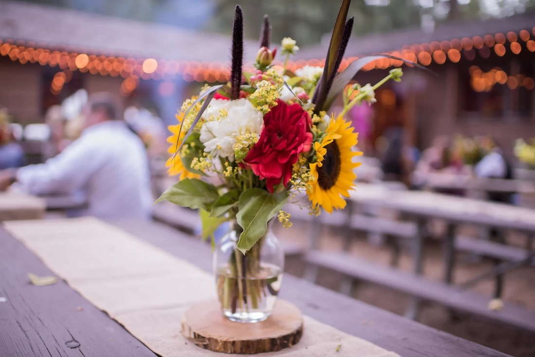 Silver Falls Weddings offer a rustic state park setting that is private and quiet. Pictured here are summer flowers picked and displayed in a vase sitting on a pice of wood. Lights twinkle out of focus in the distance. Guests sit at tables and talk.