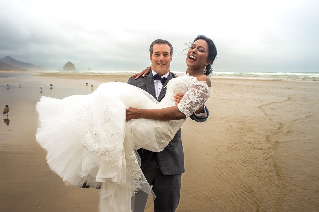 Photographers Portland at a Trash The Dress in the Rain on the Beach, man carries his bride in a white dress as a wave splashes under them. they are standing in the ocean up to their ankles, it is raining, seagulls stand very close not bother by us. the couple laughs at the situation that might make some cry. It is just beautiful
