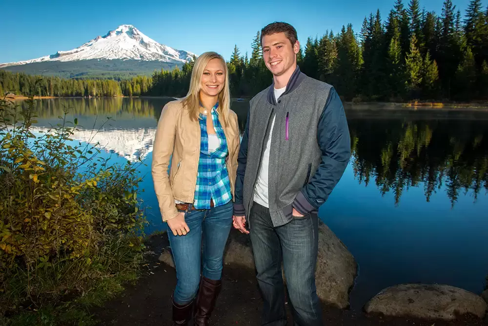 A couple stands with jackets on facing the camera with smiles, A mountain and trees reflect in a lake behind them. Unforgettable Moment - Mountain Engagement Photos 
with
​ Photojournalist Photographer Robert Knapp