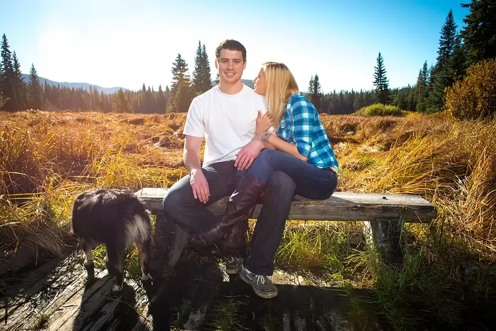 a couple sit on a bench in the mountains in a meadow the lady snuggles on the shoulder of the man, a dog is hanging around their feet Unforgettable Moment - Mountain Engagement Photos 
with
​ Photojournalist Photographer Robert Knapp