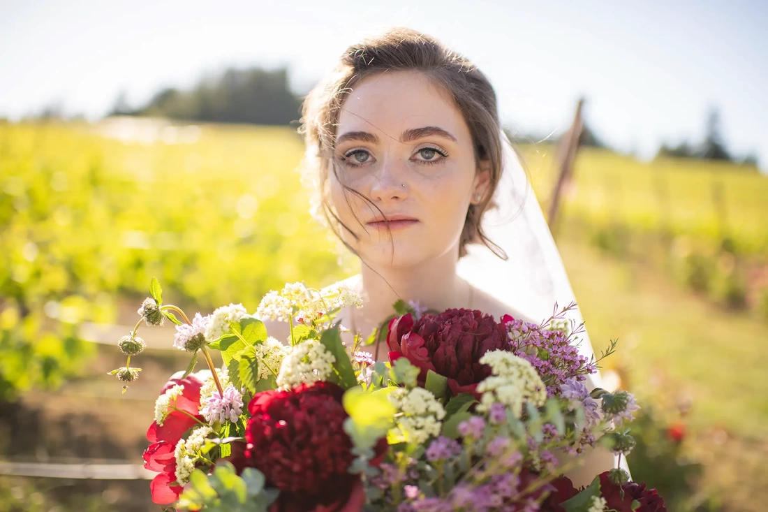 ​Vineyard Wedding Oregon where a bride stands with her bouquet in the sun with the grape vines behind her 