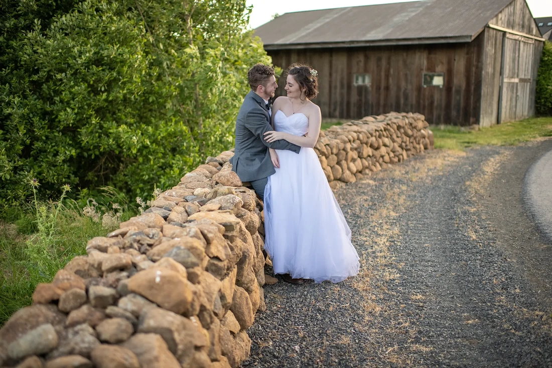 ​Vineyard Wedding Oregon with a bride and groom sitting on a stone wall next to a barn 