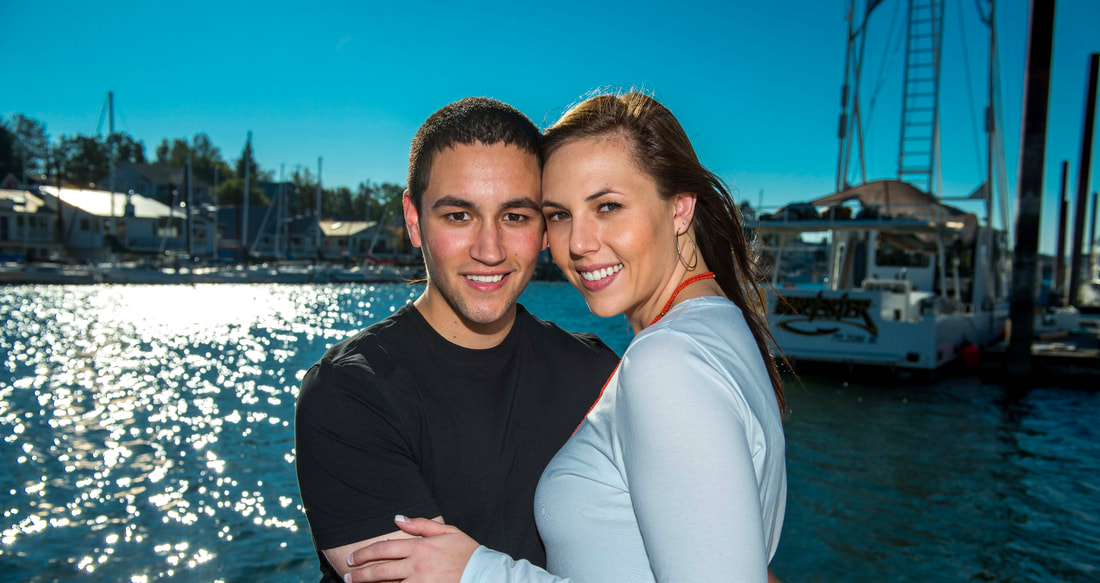 Engagement photo, a couple stands on the dock cheek to cheek looking at the camera. Sailboats are moored in the background