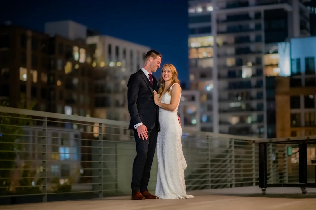 Wedding Photographers in Portland  at the EcoTrust Robert Knapp Photographer A bride and groom stand on top of a rooftop in portland at night. They are sharply in focus, the lights of the city behind them are out of focus. They are very happy to be married.