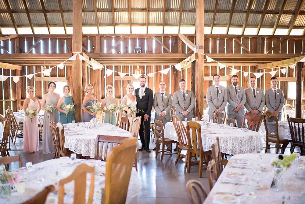 Tin Roof Weddings Barn Weddings Venues Near Me from Photographer Robert Knapp Wedding party inside the barn the full wedding party is all facing the camera. The light shines through the walls and boards of the barn.