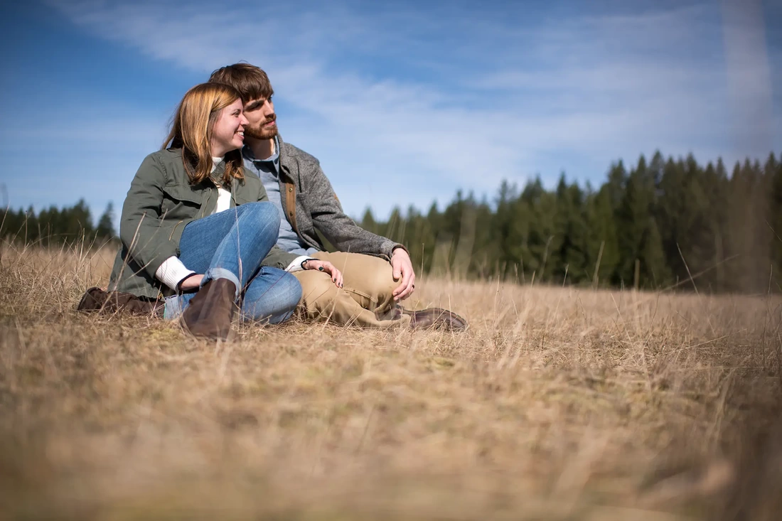 ​Winter Waterfall Engagement Photos from Photographer Robert Knapp a couple sits in a field of dry brown grass und a rich blue sky. The couple nuzzles temples 