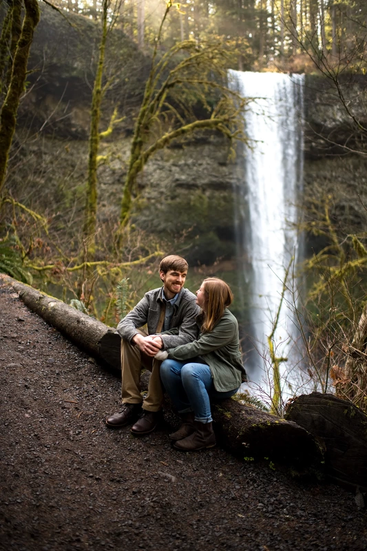​Winter Waterfall Engagement Photos from Photographer Robert Knapp a couple sits on a log along a path to a waterfall. 