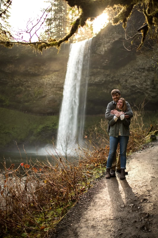​Winter Waterfall Engagement Photos from Photographer Robert Knapp a couple holds each other in a ray of sunlight shining down from above a waterfall during an engagement photos shoot 